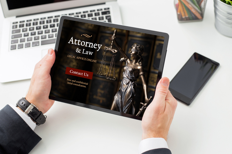 A person holds a tablet displaying a law firm website with the words ‘Attorney & Law – Legal Advice Online’ and a red ‘Contact Us’ button. The screen also shows a bronze statue of Lady Justice holding scales, with a blurred laptop and phone in the background.